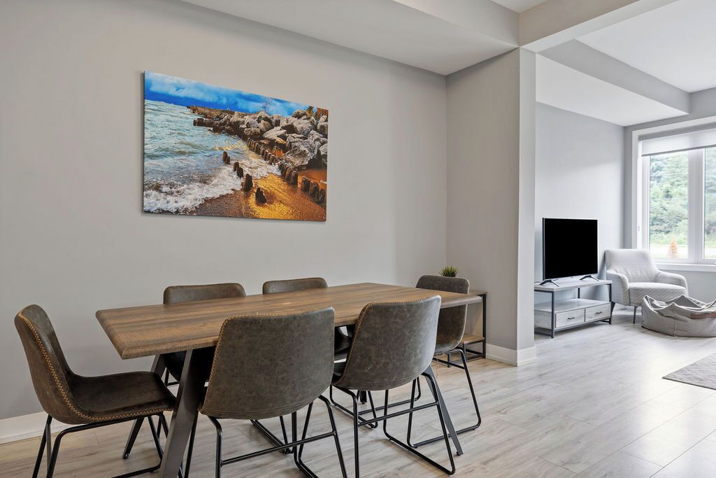 Un espace repas moderne avec une table et des chaises en bois, un tableau représentant un paysage côtier et un téléviseur en arrière-plan.
