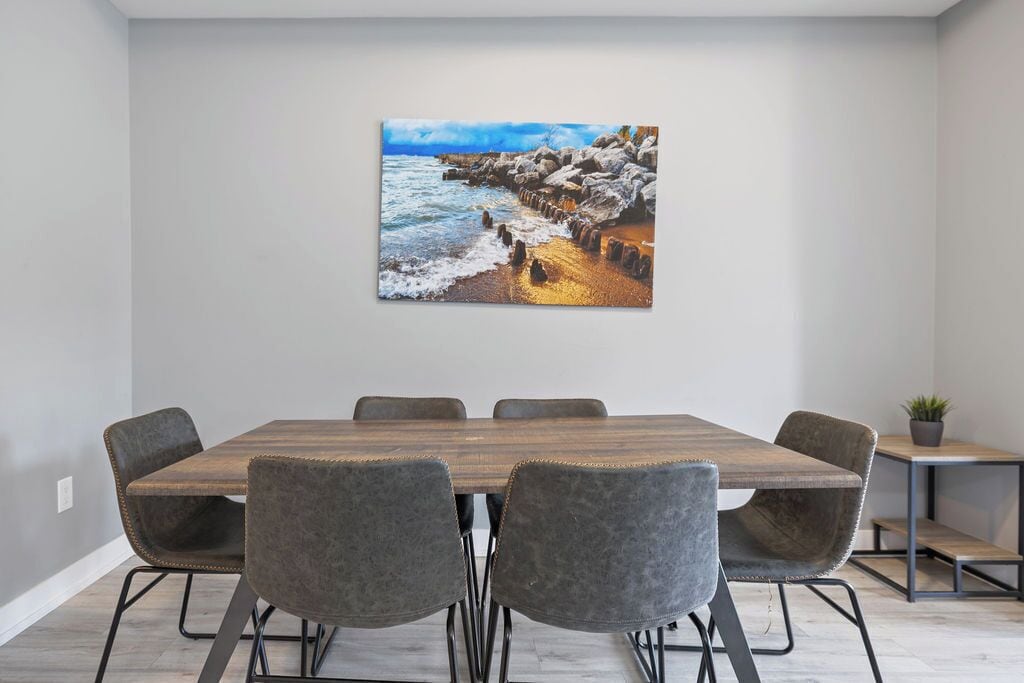 Un comedor moderno con una mesa de madera y sillas grises tapizadas, un cuadro de una costa rocosa en la pared y una pequeña planta en maceta sobre una mesita auxiliar.