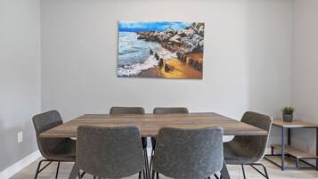 Un comedor moderno con una mesa de madera y sillas grises tapizadas, un cuadro de una costa rocosa en la pared y una pequeña planta en maceta sobre una mesita auxiliar.