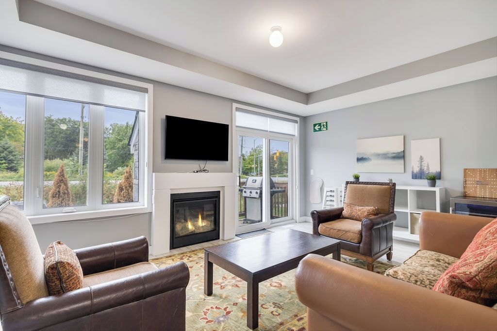 A spacious living room with leather sofas, a fireplace, a flat-screen TV, and large windows offering a view of the outdoors.