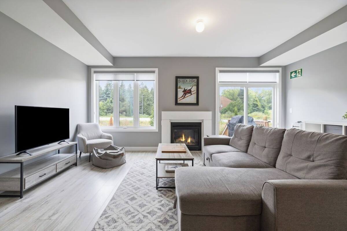 A modern living room with a flat-screen TV, a fireplace, a sofa, and large windows offering a view of greenery.
