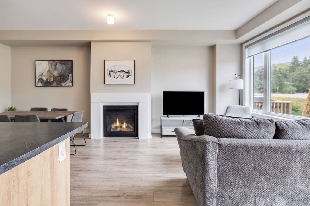 A modern living room with a fireplace, a flat-screen TV, and a large window offering a view of the outdoors.