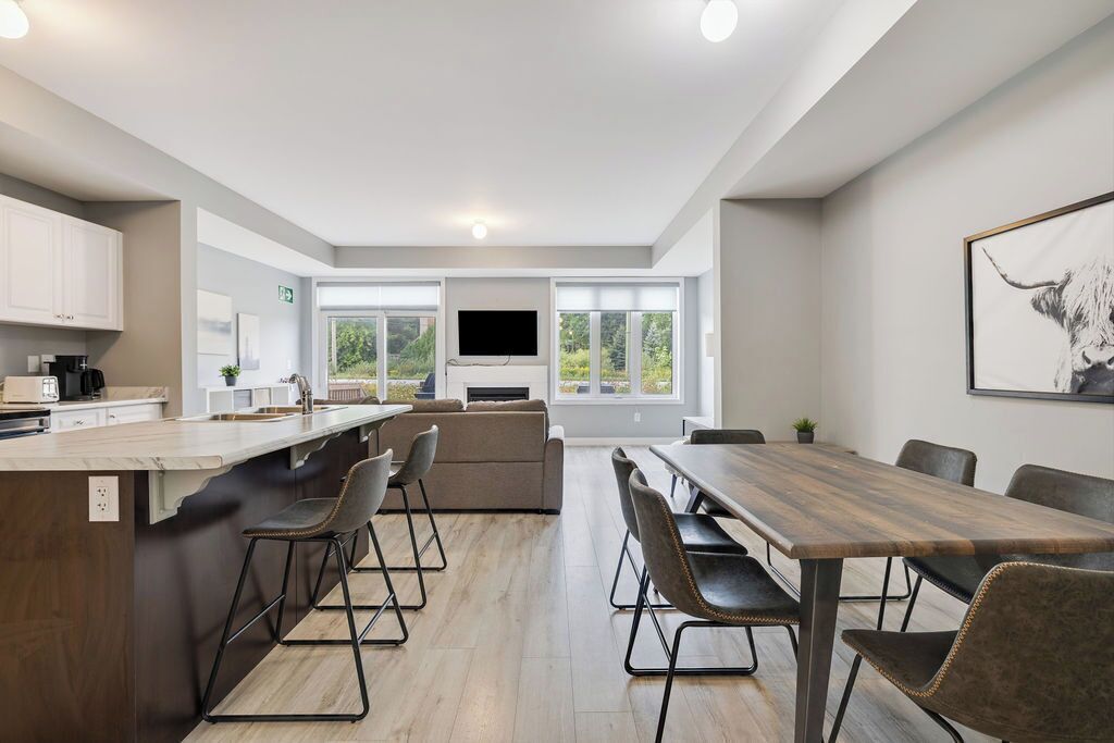 A modern kitchen with a central island, dining area with a long table and chairs, and a large window providing a view of greenery.