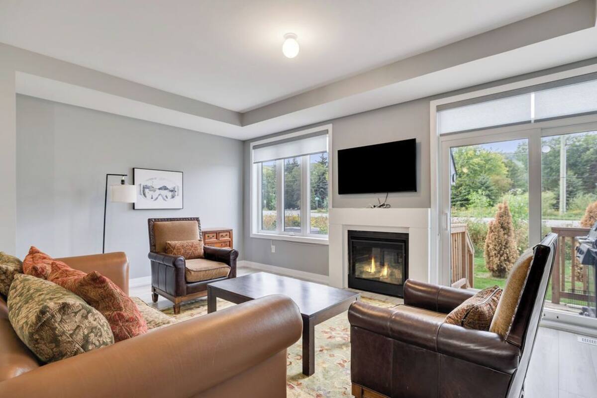 A spacious living room with a fireplace, leather sofas, a coffee table, and large windows offering a view of the outdoors.