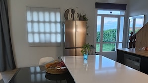 A modern kitchen with a large island, a refrigerator, and a view of greenery outside.