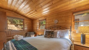Camera da letto in una baita di legno, con un letto, un comodino, una lampada e una finestra vista alberi.