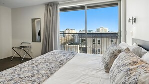 A hotel room with a large bed, a balcony with a city view, and a chair by the window.
