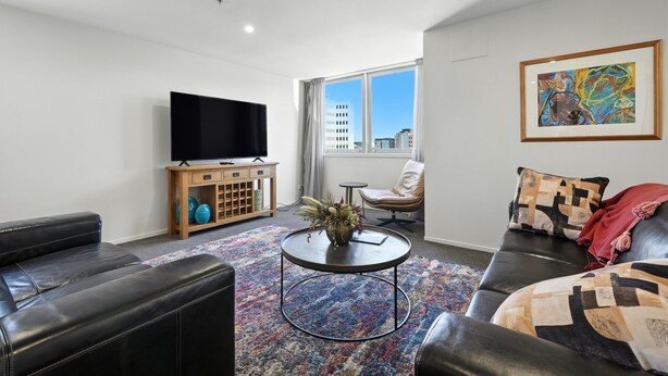 A modern living room with a flat-screen TV, leather sofas, a coffee table, and a large window with a city view.
