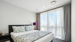 A hotel room with a large bed, a nightstand, a lamp, and a view of the city through sheer curtains.