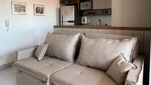 A modern living room with a beige sofa, a kitchen area with a microwave and cabinets, and two framed pictures on the wall. - Red  (Bauru)