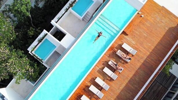 An aerial view of a modern hotel with a rooftop swimming pool, multiple lounge areas, and a wooden deck.