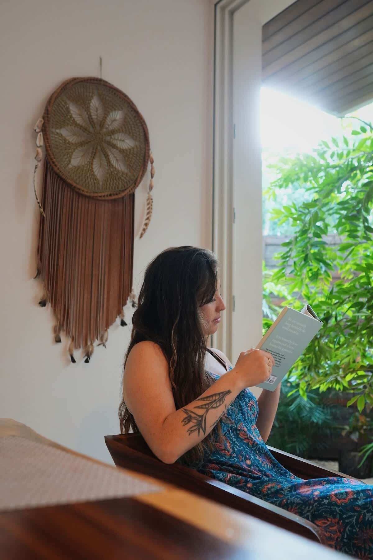 A person reading a book in a room with a wall hanging and a window with a view of greenery.