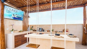 A lounge area with a wooden ceiling, a large TV screen displaying 'LOUNGE COVERS POPULAR SONGS', a kitchen counter with various items, and a wooden bench. - 3 Bedrooms Penthouse-Private Pool-Free parking (Tulum)