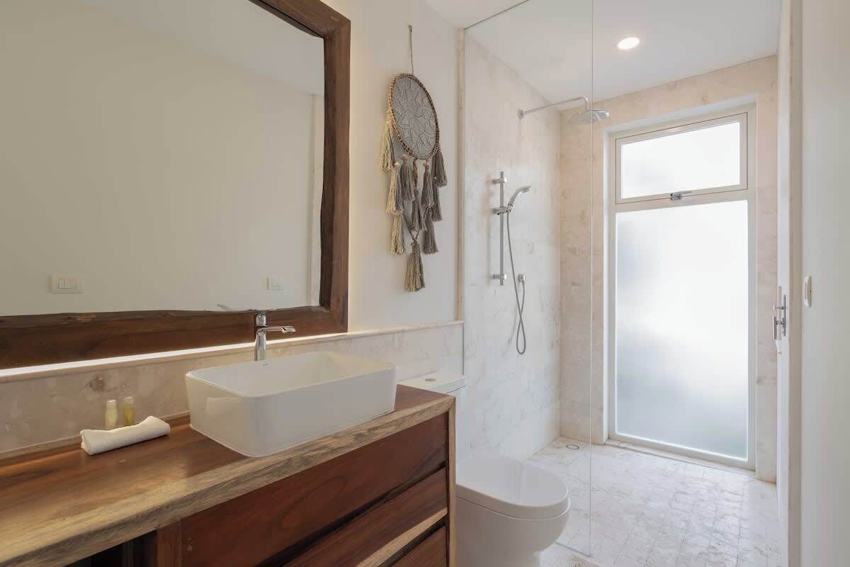 A modern bathroom with a large mirror, a wooden vanity, a white sink, a dreamcatcher, a shower, and a frosted glass door.