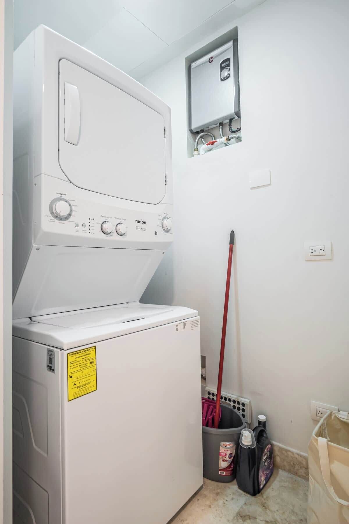 A laundry area with a washing machine and dryer stacked vertically, a mop, cleaning supplies, and a small electrical panel on the wall.