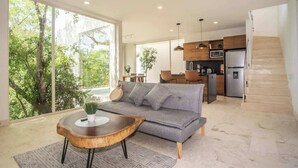 En moderne stue med en grå sofa, et træsofabord og store vinduer med udsigt til grøn vegetation.