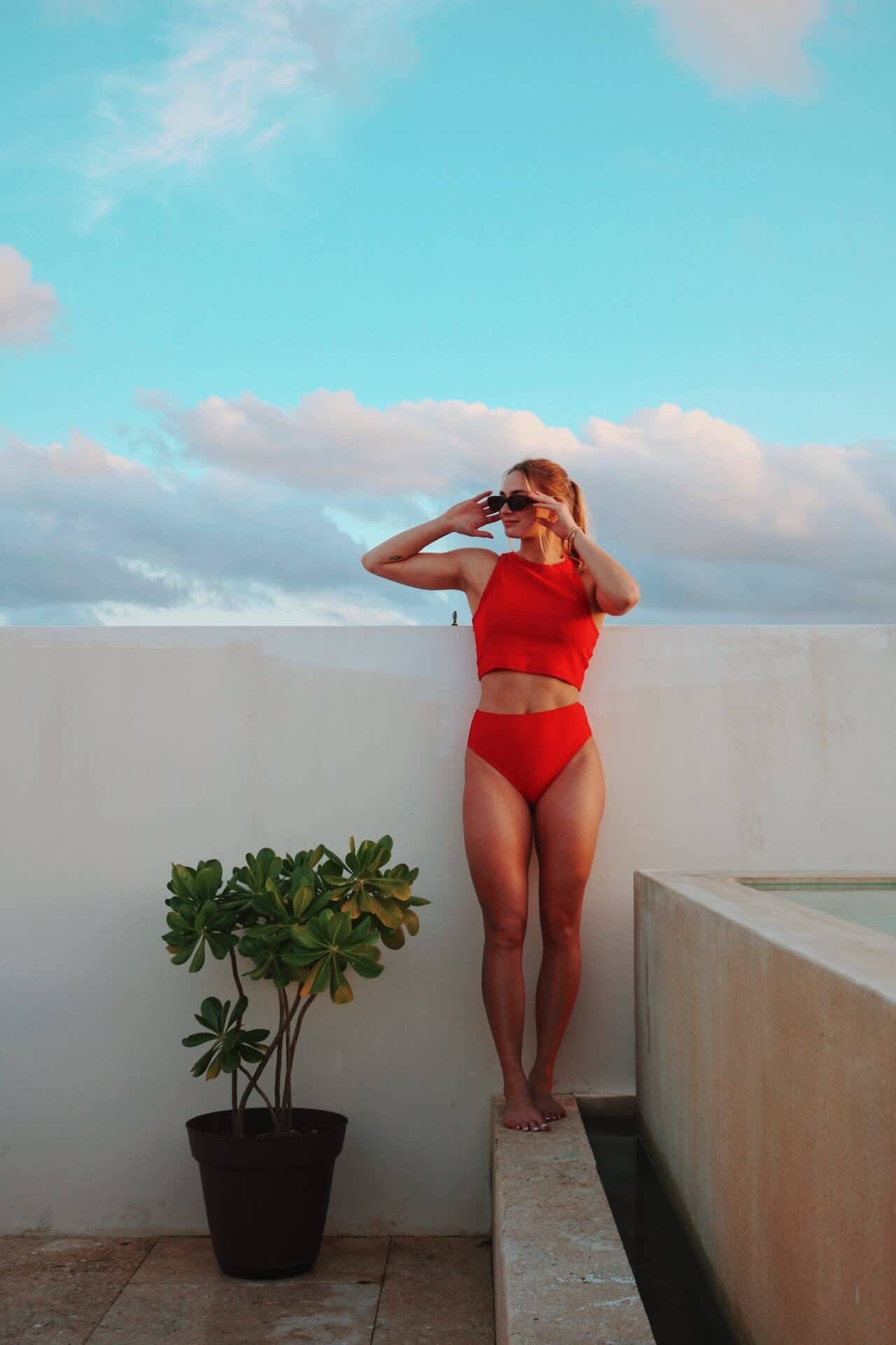A person standing on a balcony wearing a red swimsuit, with a potted plant and a clear sky in the background.