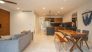 A modern living room with a sofa, dining table, and chairs. There is a kitchen area in the background with cabinets and appliances.