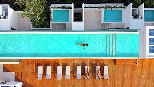 An aerial view of a modern swimming pool with a person swimming, wooden deck with lounge chairs, and a white building with a blue-tiled roof.