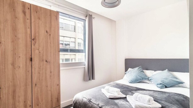 A bedroom with a bed, a wardrobe, a window with curtains, and a view of buildings outside.