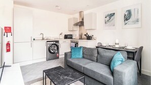 A modern apartment with a grey sofa, a small dining table, a kitchen with a microwave, and a washing machine.