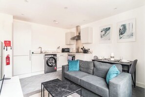 Un apartamento moderno con un sofá gris, una pequeña mesa de comedor, una cocina con microondas y lavadora.
