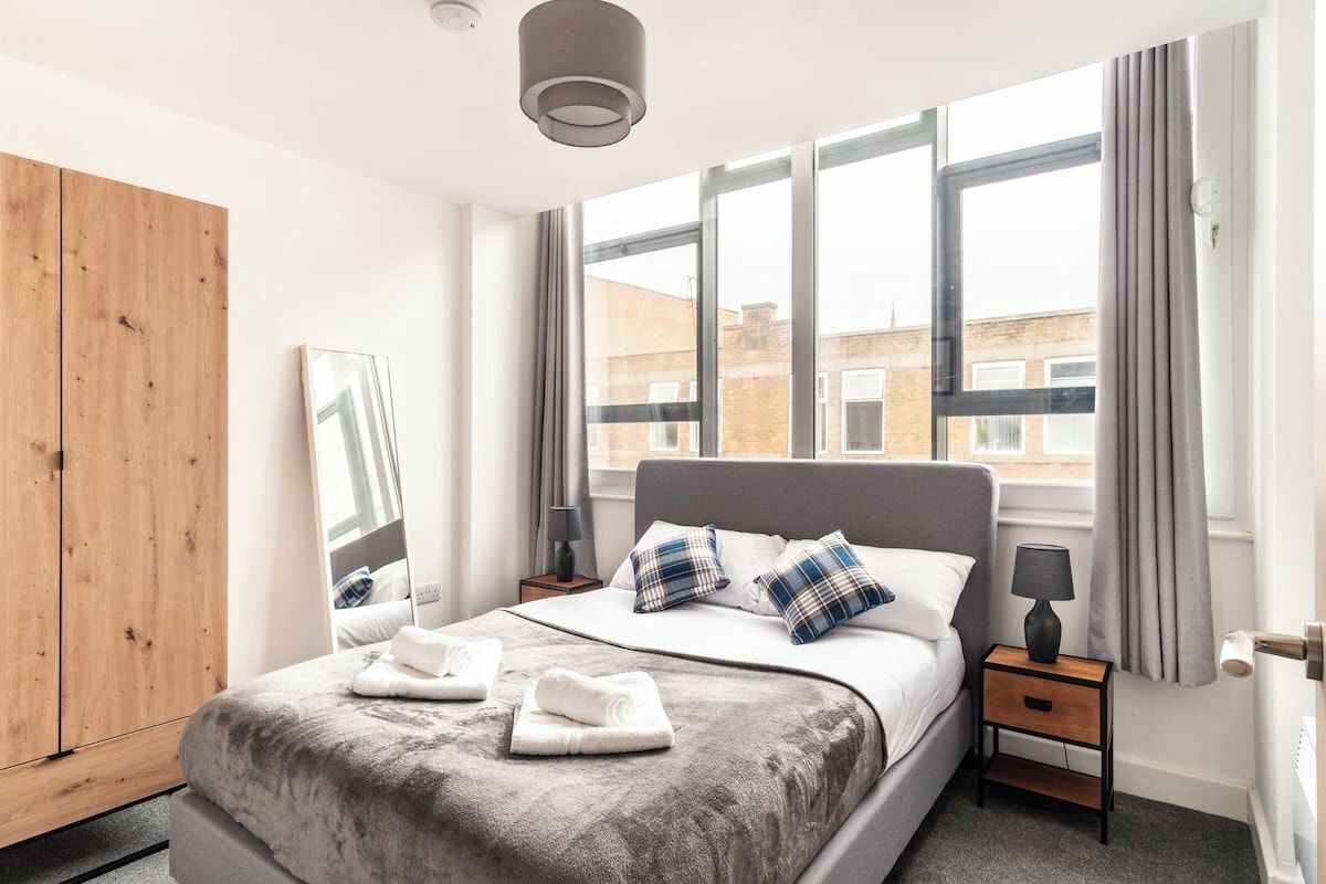 Habitación de hotel moderna con una cama grande, un armario de madera, una mesita de noche con lámpara y vistas a la ciudad a través de amplios ventanales.