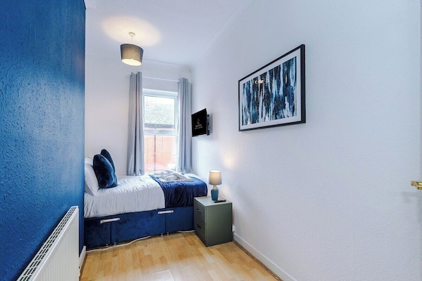 A bedroom with a bed, bedside table, lamp, window, and artwork on the wall.