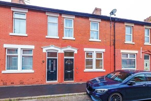 A red brick terraced house with a black front door, white-framed windows, and a satellite dish on the roof.