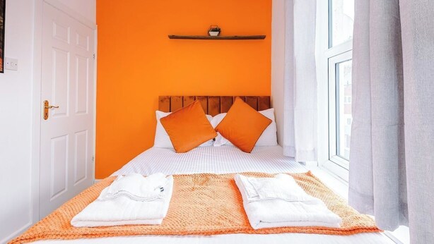 A hotel room with a bed, pillows, a shelf with a small item, and a window with curtains. - Preston PR2 Home | Ashton-on-Ribble | Parking (Lancashire)