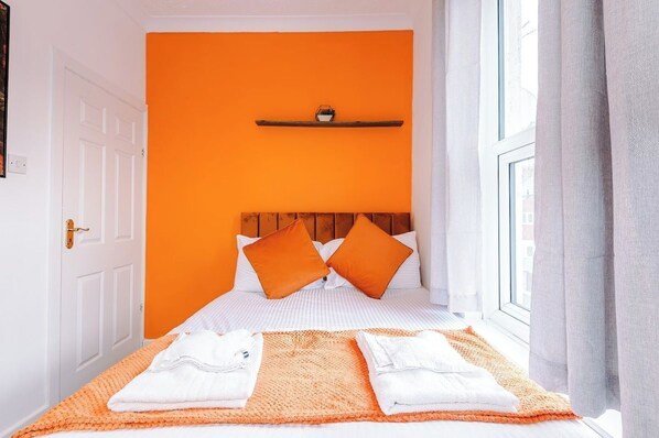 A hotel room with a bed, pillows, a shelf with a small item, and a window with curtains.