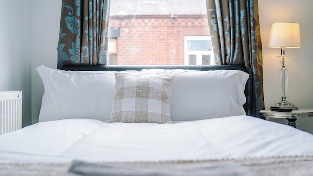 A neatly made bed with white linens, a checkered throw pillow, and a lamp on a bedside table. - 15%OFF|Stay7PayLess|Business|Parking|WiFi|Sleeps7 (Lancashire)