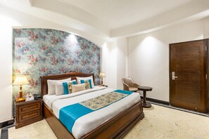 Una habitación de hotel con una cama grande, cabecera de madera, papel tapiz floral, mesita de noche con lámpara, una silla y una puerta.
