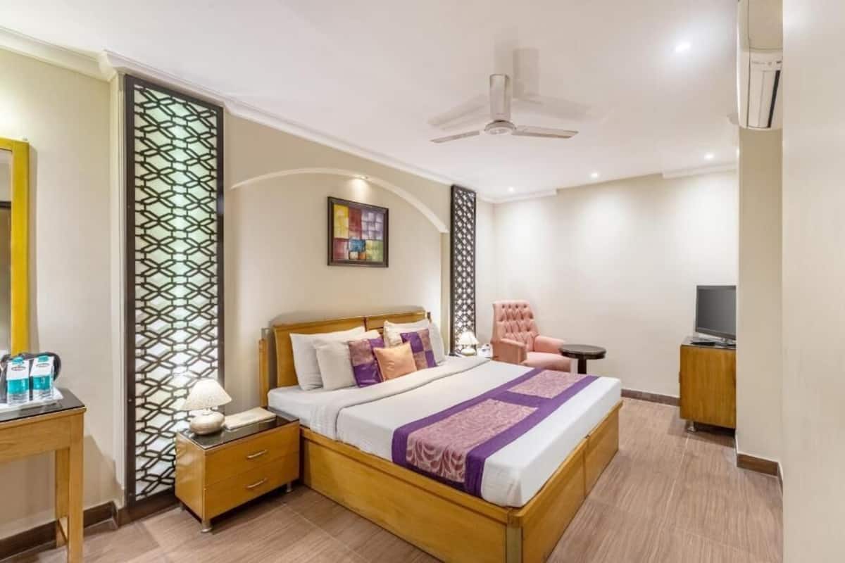 Quarto de hotel com cama, mesa de cabeceira, abajur, painel decorativo na parede, ventilador de teto e televisão sobre um móvel de madeira.