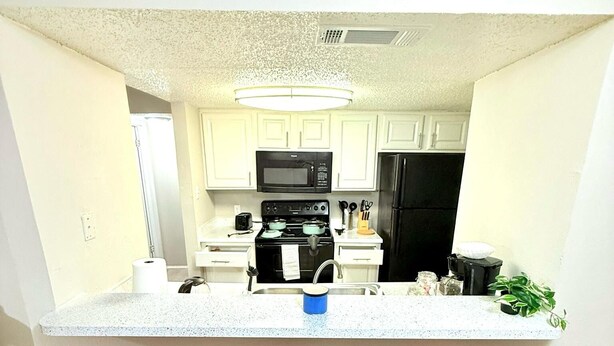 A modern kitchen with white cabinets, a black microwave, a stove, and a black refrigerator. There are four brown bar stools with metal bases.