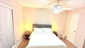 A bedroom with a bed, a ceiling fan, two bedside lamps, a window with blinds, and a door.