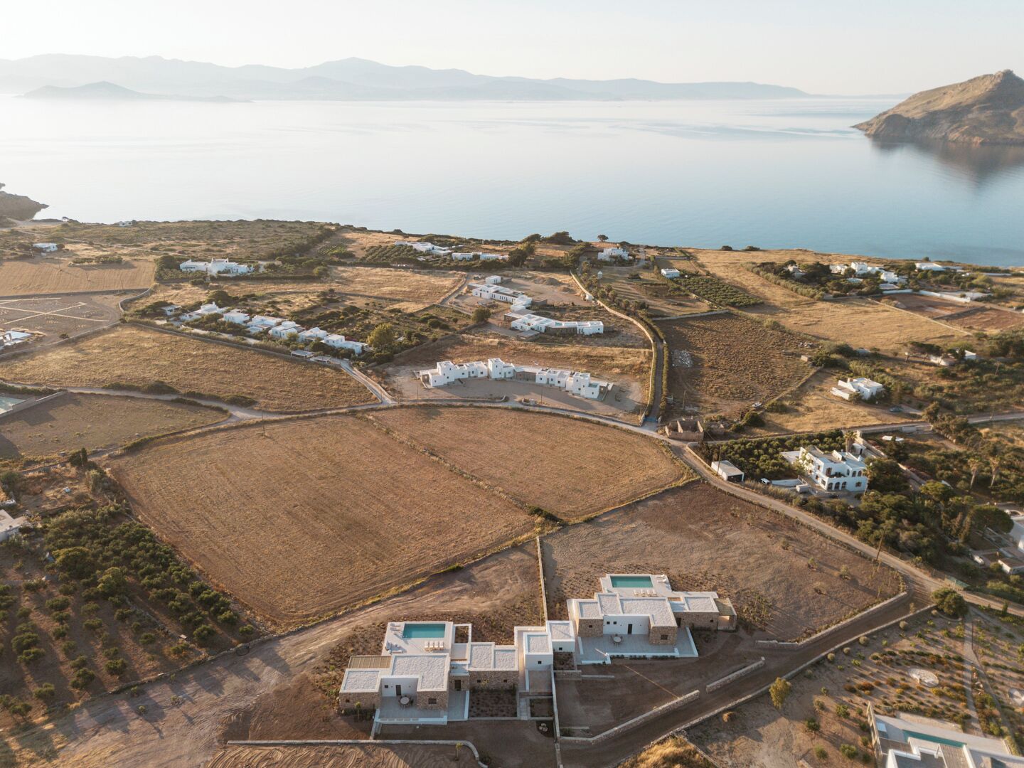 An aerial view of a coastal area with white buildings, including a cluster with a pool, surrounded by fields and a body of water.