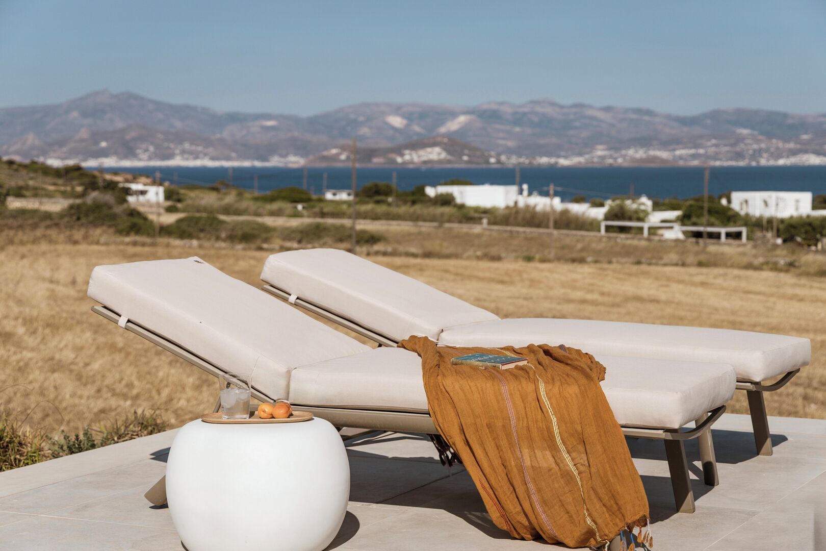 Two white lounge chairs with a brown cloth draped over one, a white vase, and a view of the sea and mountains in the background.