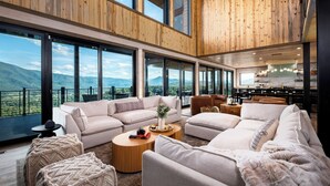 A spacious living area with a large window overlooking a mountainous landscape, furnished with sofas, a coffee table, and a kitchen in the background. - LUXURY NEW CONSTRUCTION! Mountain Horizon–Pool, Spa, Sports Court, Golf, Views (Pigeon Forge)