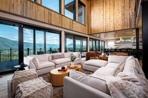 A spacious living area with a large window overlooking a mountainous landscape, furnished with sofas, a coffee table, and a kitchen in the background.