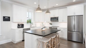 A modern kitchen with white cabinets, a stainless steel refrigerator, a microwave, and a large island with bar stools. - True North - Ocean Bliss Retreat in Kure Beach (Kure Beach)