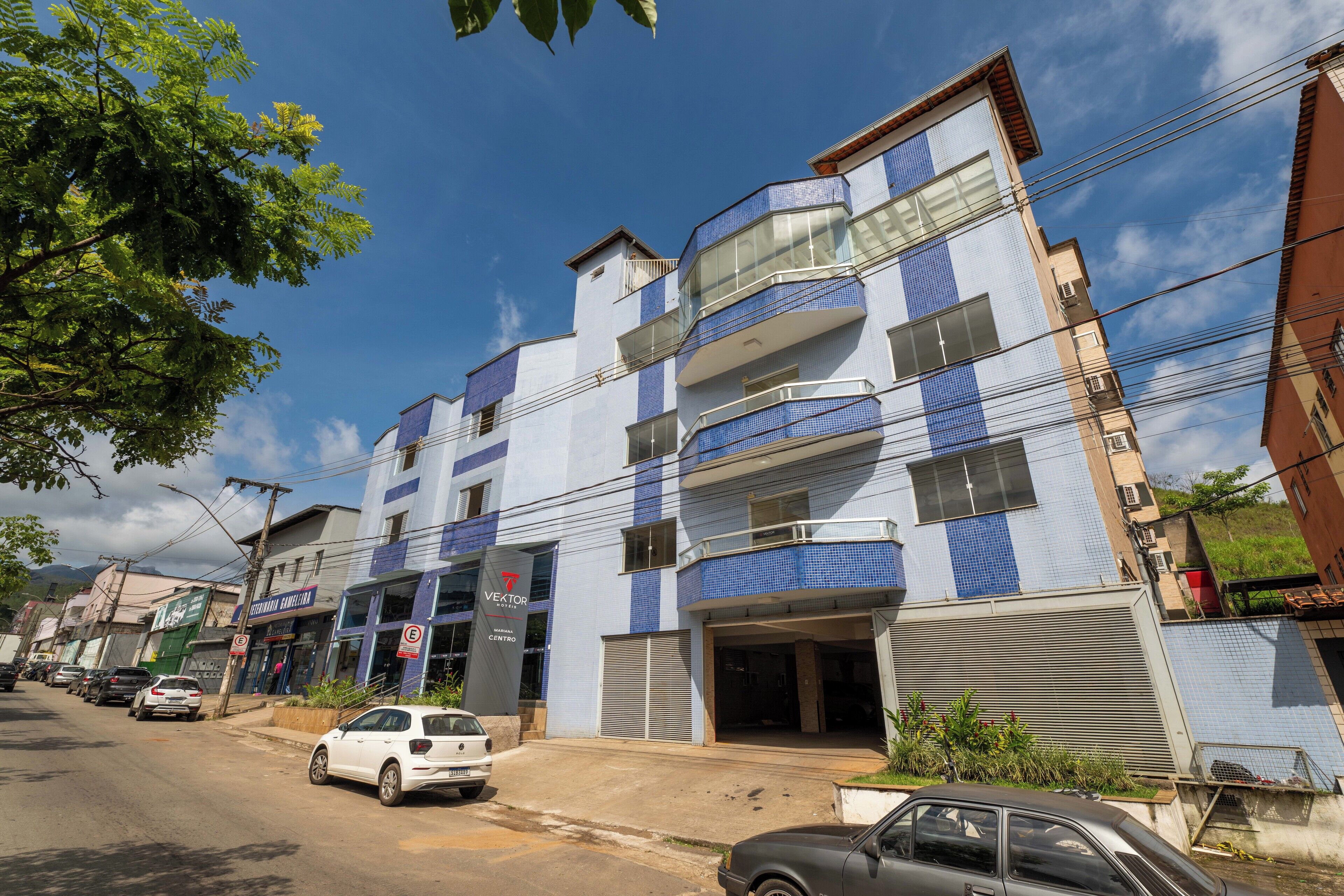 A multi-story building with a blue and white facade, a visible entrance, and a parking area with cars.