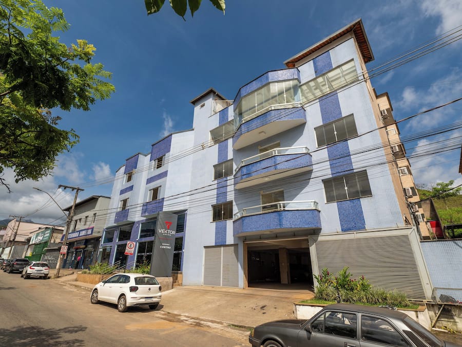 A multi-story building with a blue and white facade, a visible entrance, and a parking area with cars.