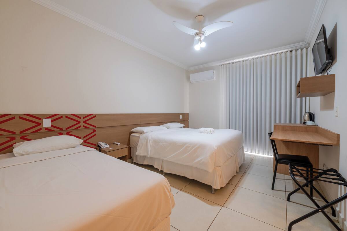 A hotel room with two beds, a desk, a chair, a TV, and a ceiling fan.