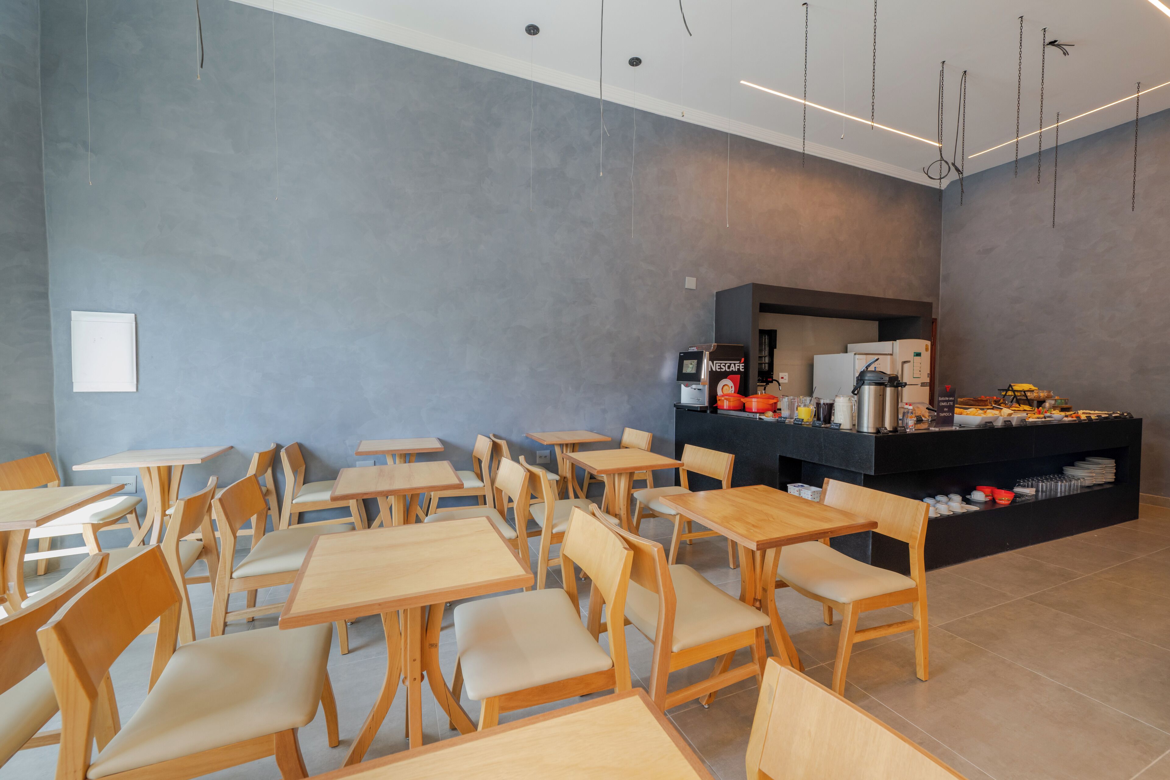 A modern cafe with wooden tables and chairs, a coffee station, and a grey textured wall.