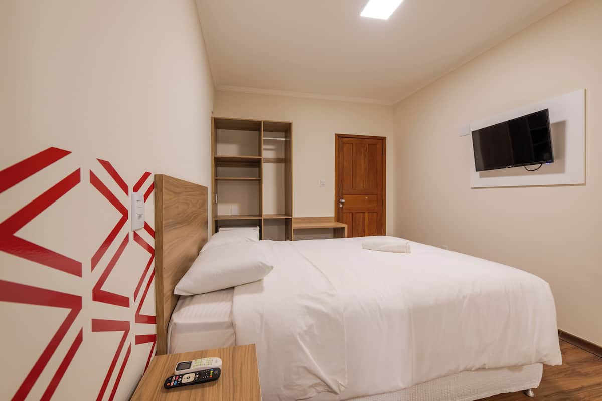 A hotel room with a bed, a TV mounted on the wall, a wooden nightstand, and a wardrobe.