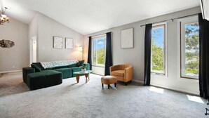 A spacious living room with a green sofa, a small round table, and a chair. There are large windows with curtains, a chandelier, and framed artwork on the walls. - Stylish 2BR Loft, Near ISU, Hospitals & Downtown (Bloomington)