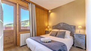 Ein Schlafzimmer mit einem hölzernen Kopfteil, einem großen Bett mit weißer und grauer Bettwäsche, zwei Nachttischen mit Lampen und Blick auf eine grüne Landschaft durch ein Fenster.