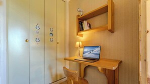 Ein Zimmer mit Schreibtisch aus Holz, einem Laptop, einem Stuhl und einem Bücherregal.