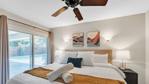Quarto de hotel com uma cama grande, um ventilador de teto, duas luminárias fixadas na parede e vista para uma piscina através de uma porta de vidro deslizante.